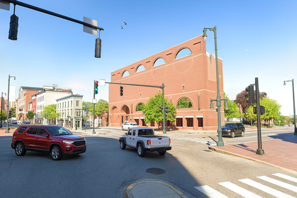 A bright, clear day at a city intersection with a large red brick building in the background. Several vehicles, including a red SUV, a white pickup truck, and a dark crossover, move through the intersection. The streets are clean and empty of pedestrians, and the surrounding buildings and traffic lights are clearly visible.