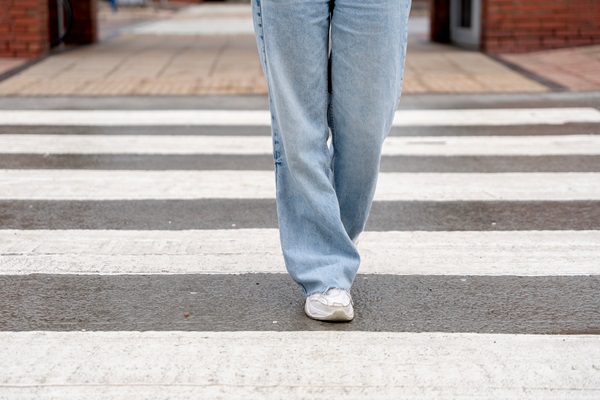 Person wearing jeans and sneakers walking across a wet crosswalk in an urban area.