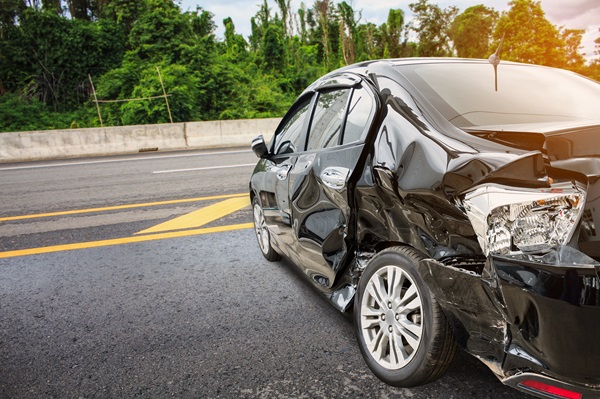 A black sedan with significant damage to the rear and side parked on the shoulder of a highway next to a dense forest in Maine.