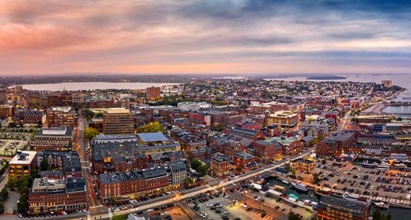 An aerial view of a Portland, Maine, at dusk, showing densely packed brick buildings, harbor docks with boats, and a sunset sky over the horizon.