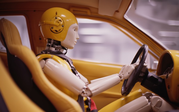 A side profile of a yellow crash test dummy seated in a vehicle, wearing a matching yellow helmet and secured by a seatbelt behind the steering wheel.
