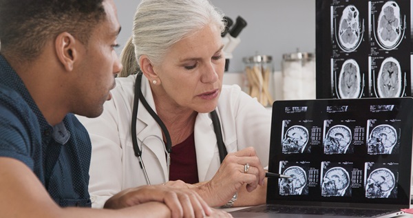 A doctor in a white coat sits with a patient, using a pen to point out specific areas on a series of brain scans displayed on a laptop screen to explain a concussion diagnosis.